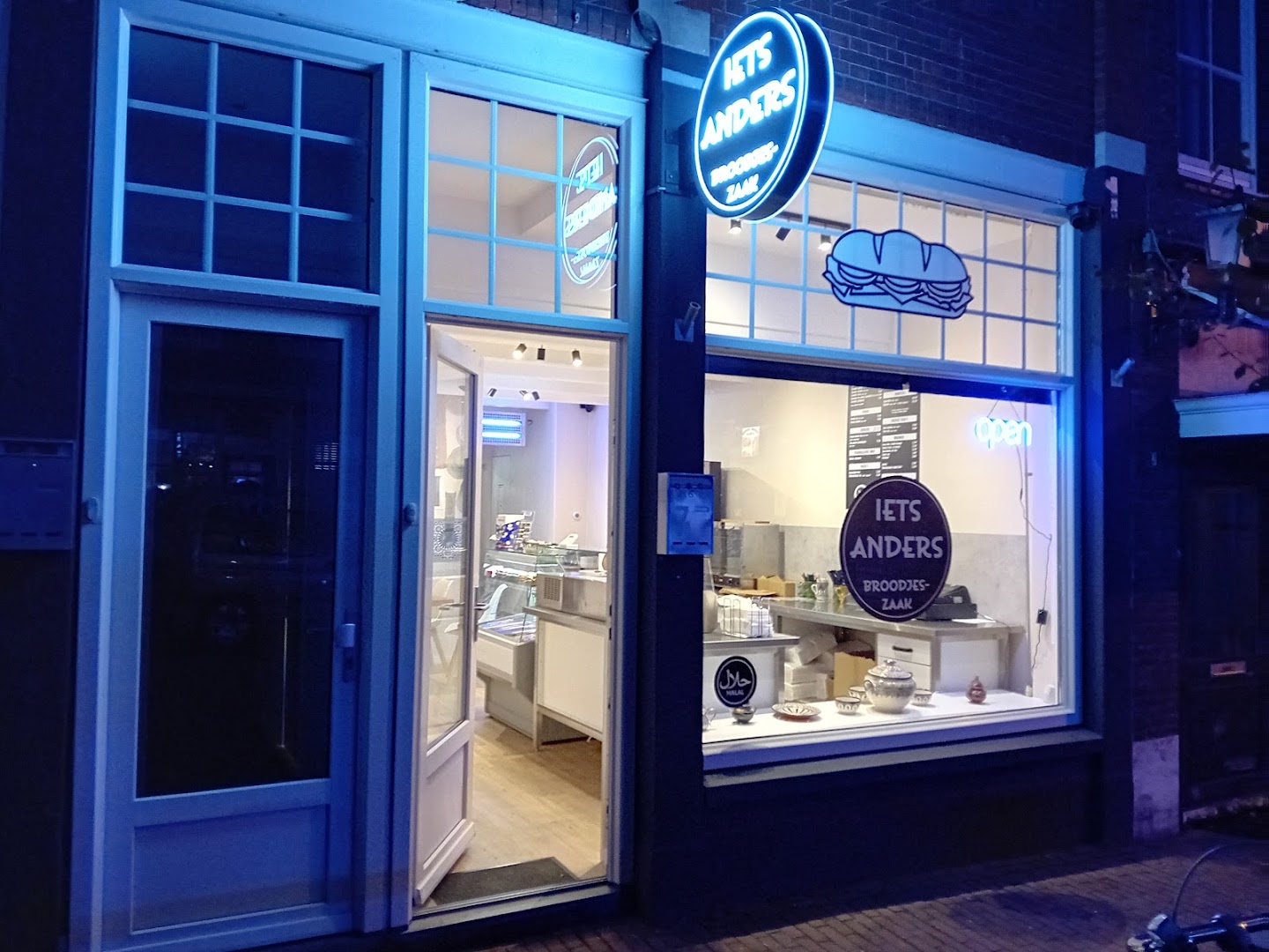De gevel van Iets Anders Broodjeszaak in Oudenbosch bij nacht, met het echte blauwe neonbord en neon-sandwich